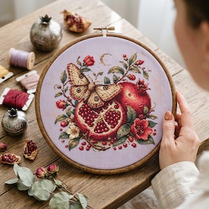 Peut inclure: Un cercle de broderie au point de croix avec un motif détaillé d'un papillon, de grenades et de fleurs. La broderie est réalisée sur un tissu violet clair, encadrée par un cercle en bois. La palette de couleurs comprend des rouges, des verts et des bruns.