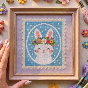 Könnte beinhalten: Kreuzstichkunst eines weißen Hasen mit Blumenkranz, gerahmt in einem hellen Holzrahmen. Der Hase ist vor einem blauen Hintergrund mit weißen Punkten platziert, der einer Briefmarke ähnelt. Dekorative Blumen und Stickgarn umgeben das Kunstwerk.