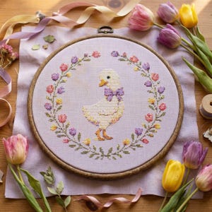 May include: A cross-stitch embroidery hoop featuring a yellow duck with a purple bow tie, encircled by a floral wreath of pink, purple, and yellow flowers. The embroidery is on lavender fabric, with tulips and ribbons around the hoop.
