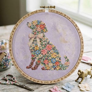 Puede incluir: Bordado de punto de cruz de la silueta de un perro lleno de flores coloridas, en un aro de madera. El perro tiene un lazo y está rodeado de un fondo lila claro. El aro de madera está decorado con diseños florales y de huesos.