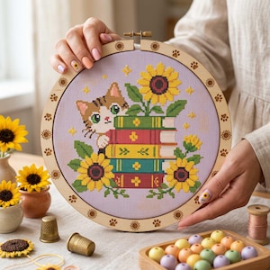 May include: A circular wooden embroidery hoop frame featuring a cross-stitch design of a brown and white cat peeking over a stack of four colorful books, surrounded by yellow sunflowers and small gold stars on a light purple fabric background.