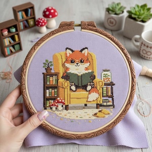 Peut inclure: Broderie au point de croix dans un cercle en bois représentant un renard de dessin animé lisant dans un fauteuil jaune. Le renard a une fourrure orange et blanche. Des étagères, un champignon et une plante sont également dans la scène. Le fond est en tissu violet clair.