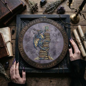 May include: Cross-stitch embroidery of a blue and yellow dragon perched atop a stack of books, holding a book. The embroidery is set in a wooden hoop with a dark frame, featuring runes. The background is a muted purple.
