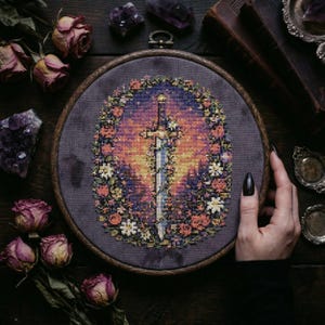 Puede incluir: Bordado de punto de cruz en un aro de madera con un diseño de espada. La espada está centrada sobre un fondo de color atardecer, rodeada por una corona floral. El bordado está sobre una tela gris oscura.