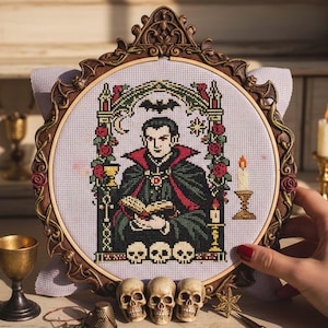 May include: A framed cross-stitch featuring a vampire design. The artwork depicts a vampire in a black and red cape, holding a book, surrounded by roses, skulls, and a bat. The frame is ornate, with a brown finish. A candle and chalice are also visible.