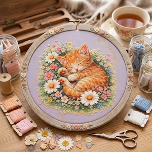 Könnte beinhalten: Kreuzstich-Stickerei eines schlafenden, orangefarbenen Kätzchens, umgeben von bunten Blumen, in einem Holzrahmen mit floralen Details. Garnrollen, eine Schere und eine Tasse Tee sind ebenfalls zu sehen.