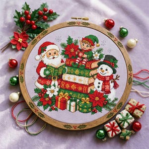May include: A circular wooden embroidery hoop containing a cross-stitch design of Santa Claus, an elf, and a snowman surrounded by stacked books, poinsettias, and wrapped gifts on a light purple fabric background with holly leaf engravings.