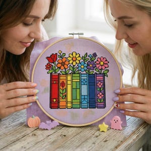 May include: A circular wooden embroidery hoop containing a cross-stitch pattern on light purple fabric. The design features a row of colorful books in rainbow order, topped with various pixelated flowers in red, orange, yellow, blue, and pink.