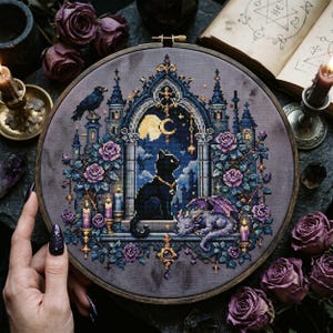 May include: A circular embroidery hoop containing a cross-stitch design on grey fabric. The image features a black cat sitting in a gothic archway with a small dragon, purple roses, a raven, candles, and a crescent moon against a night sky.