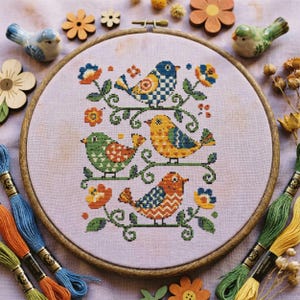 May include: A cross-stitch embroidery featuring five colorful birds perched on a leafy branch, framed in a wooden hoop. The birds have intricate patterns in blue, green, orange, and yellow. Embroidery floss in various colors and wooden flower accents surround the hoop.