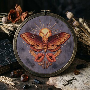May include: A cross-stitch design of a moth with a skull-like head and sunburst detail, in shades of brown and orange, set in a round, bronze-colored frame. The design includes wheat and poppies. The background is a muted gray fabric.