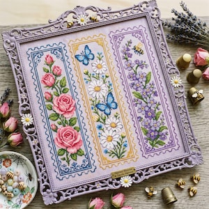 May include: A square lavender frame containing three vertical embroidered panels featuring pink roses, white daisies with blue butterflies, and purple flowers with a bee. The frame is decorated with small daisies and bees, with a metal plaque reading Harvest Blooms & Gentle Harvest.
