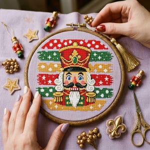 May include: A circular wooden embroidery hoop containing a cross-stitch pattern of a nutcracker soldier. The nutcracker wears a red and gold uniform with a tall hat, set against a background of horizontal red, green, and yellow brushstroke stripes.