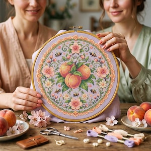 May include: A finished cross-stitch embroidery hoop with a peach design, flowers, and bees. The design is on a lavender background and framed in a wooden hoop. Peaches and embroidery supplies are on a wooden table.