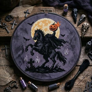 May include: A circular embroidery hoop containing a cross-stitch design of a headless horseman riding a black horse against a large yellow full moon. The scene includes a pumpkin head, flying bats, small white ghosts, and a tombstone on dark fabric.