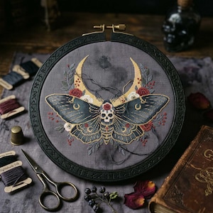 Può includere: Un ricamo a punto croce in un telaio nero raffigura una falena con un teschio sulla testa, su sfondo grigio. La falena ha ali blu scuro con dettagli dorati, circondata da lune crescenti e fiori rossi e bianchi. I materiali di cucito circondano il ricamo.