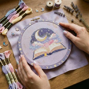 Könnte beinhalten: Ein Stickrahmen mit einem himmlischen Design, einschließlich Mond, Sterne und einem offenen Buch. Die Stickerei befindet sich auf lavendelfarbenem Stoff, mit pastellfarbenem Stickgarn und kleinen Charms.