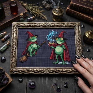 May include: Cross-stitch art in a gold frame featuring two green frog witches wearing red capes and hats. One frog holds a broom, the other a smoking chalice. The background is dark blue.