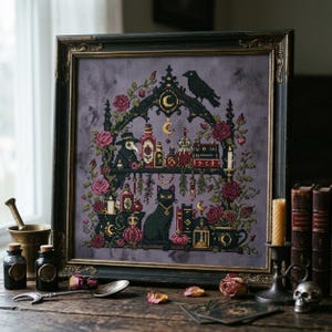 May include: A framed cross-stitch embroidery on a mottled purple fabric featuring a gothic archway, a black cat, a plague doctor, a raven, and various potion bottles surrounded by red roses and golden crescent moon symbols in a black frame.
