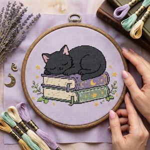 May include: Cross-stitch embroidery of a sleeping black cat on a stack of books, set in a wooden hoop. The books are decorated with moons and stars. Lavender sprigs and embroidery floss in various colors are arranged around the hoop.