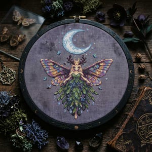 Pode incluir: Uma bordado em ponto cruz em um bastidor de madeira escura. O design apresenta uma fada com asas de borboleta, uma lua crescente e elementos florais. A paleta de cores inclui tons de roxo, verde e dourado. O bordado está sobre tecido cinza.