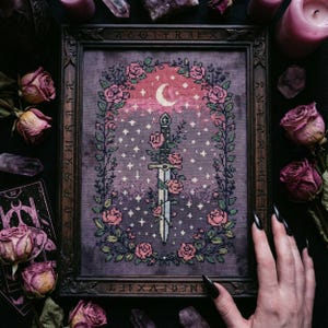 Peut inclure: Broderie au point de croix dans un cadre en bois foncé. Le motif représente une épée entrelacée de roses roses, sur fond violet étoilé avec un croissant de lune. Le cadre présente des détails sculptés et des runes.