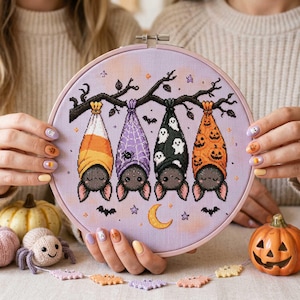 May include: A circular embroidery hoop containing a cross-stitch design of four bats hanging from a dark tree branch. The bats are wrapped in fabric patterns featuring candy corn, purple spiderwebs, white ghosts, and orange jack-o'-lanterns.