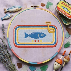 May include: A cross-stitch embroidery hoop with a blue fish design inside a sardine can. The hoop has a colourful border and is surrounded by decorative elements such as ceramic fish, sea glass, and a sardine tin.