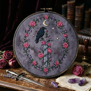 May include: Cross-stitch embroidery in a wooden hoop features a raven perched on a sword, surrounded by a wreath of pink roses and greenery. A crescent moon and stars are also included in the design. The background is a muted purple.