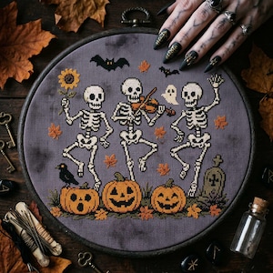 May include: A circular embroidery hoop containing a cross-stitch design on dark gray fabric. The pattern features three skeletons dancing, one playing a violin, surrounded by orange pumpkins, a sunflower, a black crow, bats, and a tombstone.