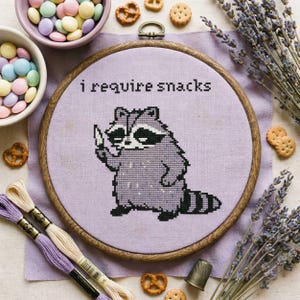 May include: A cross-stitch hoop with a raccoon design and the text "I require snacks." The raccoon is grey and black, stitched on lavender fabric. The wooden hoop is surrounded by snacks and embroidery threads.