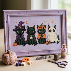 Puede incluir: Un bordado en punto de cruz enmarcado que muestra cuatro gatos negros con disfraces de Halloween: una bruja, un vampiro, una calabaza y un fantasma. El marco lavanda está decorado con pequeñas estrellas naranjas y moradas. Un frasco con la etiqueta Cackle Dust.