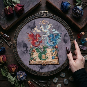 Pode incluir: Uma bordado em ponto cruz com quatro dragões em vermelho, azul, branco e verde, sobre um livro aberto. O design está emoldurado em uma moldura de madeira redonda com um fundo cinza escuro. O bordado é cercado por rosas secas, livros antigos e pequenas garrafas.