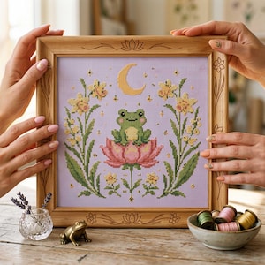 Può includere: Una cornice quadrata in legno contenente un ricamo a punto croce su tessuto viola chiaro. Il disegno raffigura una rana verde seduta su un fiore di loto rosa, circondata da narcisi gialli e una luna crescente, con motivi floreali incisi.
