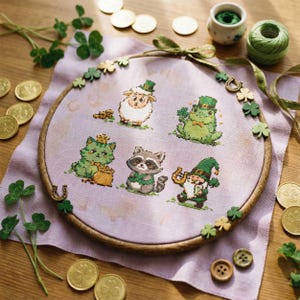 May include: A cross-stitch embroidery hoop with a St. Patrick's Day theme, showcasing a sheep, frog, cat, raccoon, and gnome, each wearing green hats and holding shamrocks. Gold coins and shamrock decorations surround the design, set on a lavender fabric, with a wooden hoop.