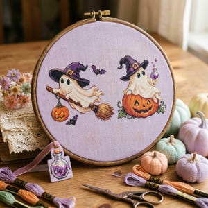 May include: A round wooden embroidery hoop containing lavender fabric with a cross-stitch design of two ghosts wearing purple witch hats, one riding a broom with a pumpkin and one sitting on a pumpkin, accompanied by small bats and a potion bottle.