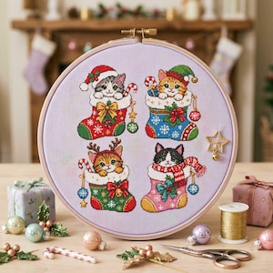 Puede incluir: Un bastidor de bordado circular con un diseño de punto de cruz de cuatro gatitos dentro de calcetines navideños. Los gatitos llevan accesorios festivos como gorros de Papá Noel y astas de reno. La tela es de color violeta claro.
