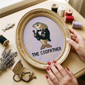 May include: An oval-framed cross-stitch embroidery featuring a fish dressed in a black tuxedo with a red rose on its lapel, holding a red rose. The text THE CODFATHER is stitched in black below the fish on a light purple fabric background.