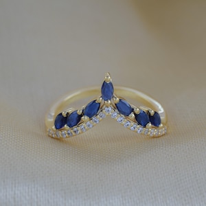 Peut inclure: Une bague chevron en or ornée d'une rangée de saphirs bleu foncé taille marquise, disposés au-dessus d'une ligne délicate de petites pierres blanches rondes et transparentes. L'anneau présente une forme en V pointue avec une finition polie.