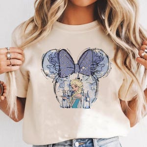 May include: Cream-colored t-shirt featuring a graphic of a cartoon character in a castle, inside a Mickey Mouse ear design. The ears are outlined in black with a blue bow with white polka dots. The character is wearing a blue dress.