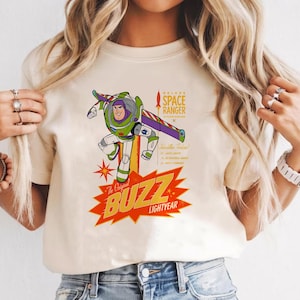 May include: Cream-colored t-shirt featuring a graphic of Buzz Lightyear in a dynamic pose. The design includes the text "The Original BUZZ LIGHTYEAR" in a red and yellow burst, and "DELUXE SPACE RANGER" with additional text. The shirt has short sleeves.