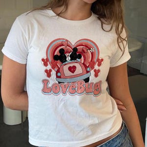 Pode incluir: T-shirt branco com um design gráfico retro. O design apresenta o texto "LoveBug", um fundo em forma de coração e Mickey e Minnie Mouse num carro. O gráfico usa cores vermelhas, rosa e azul.