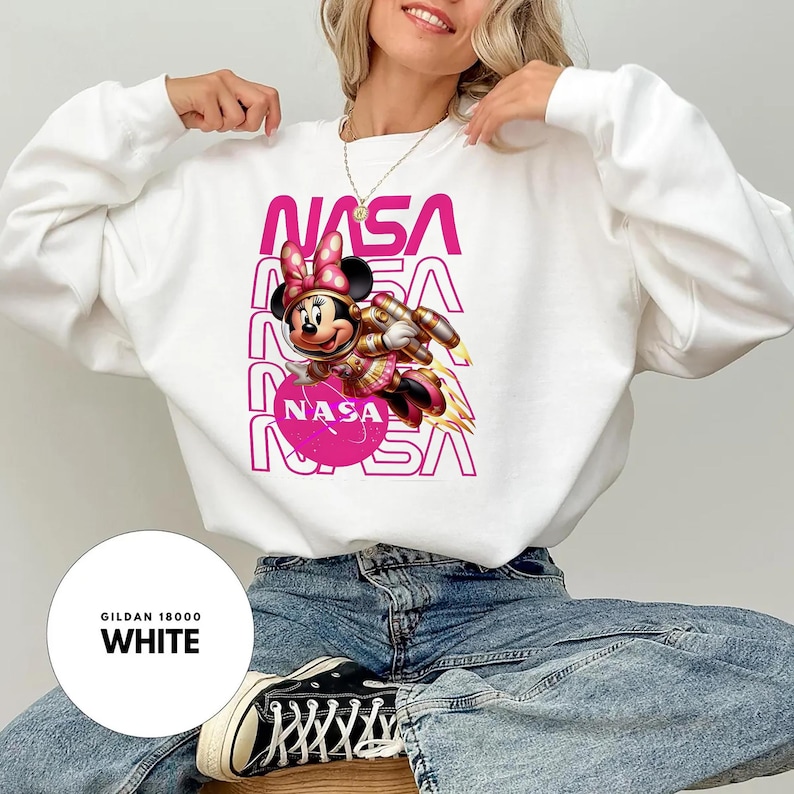 May include: White crew neck sweatshirt with a pink NASA logo and a graphic of Minnie Mouse in a space suit. The text "NASA" is repeated vertically in pink. The sweatshirt has long sleeves.