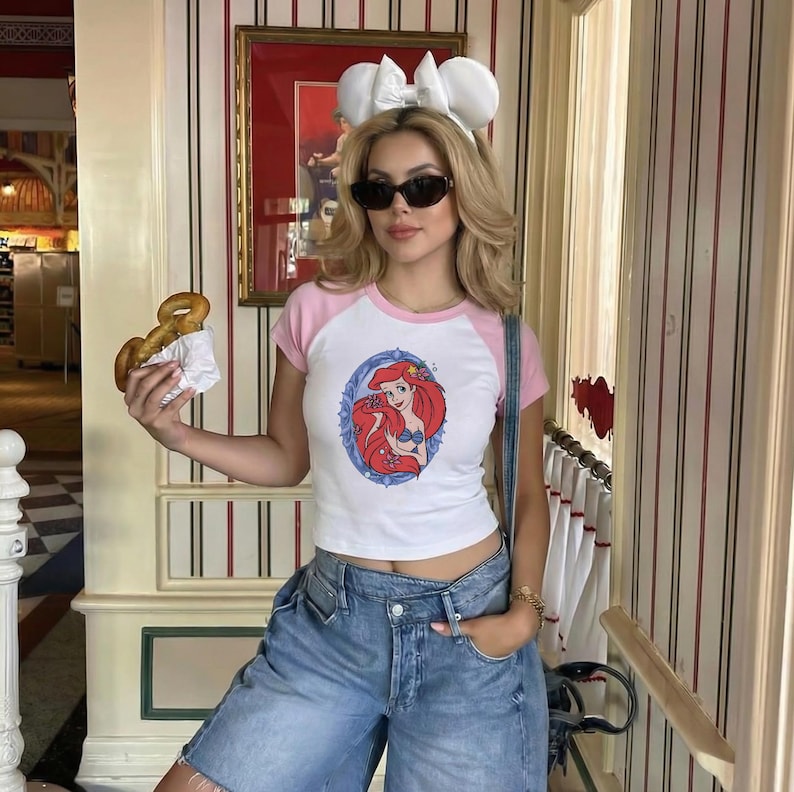 May include: White and pink ringer t-shirt featuring a graphic of Ariel from The Little Mermaid. The t-shirt is paired with denim shorts. The person is wearing sunglasses and a white bow headband.