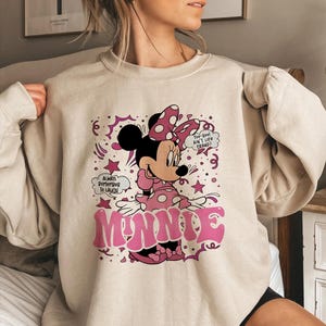Op de afbeelding: Een beige sweatshirt met een afbeelding van Minnie Mouse in roze en zwart. Minnie draagt een stippen strik en jurk. Het woord "MINNIE" staat in roze bubbelletters. Tekstballonnen zeggen "Always Remember To Laugh" en "Too Cool Ain't Life Grand?"