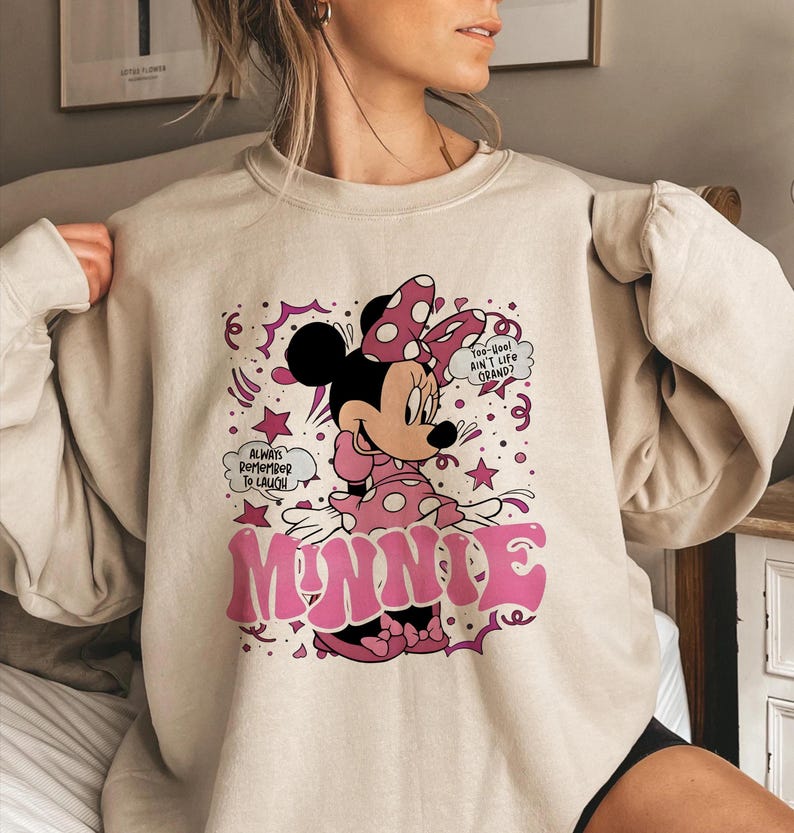 Op de afbeelding: Een beige sweatshirt met een afbeelding van Minnie Mouse in roze en zwart. Minnie draagt een stippen strik en jurk. Het woord "MINNIE" staat in roze bubbelletters. Tekstballonnen zeggen "Always Remember To Laugh" en "Too Cool Ain't Life Grand?"