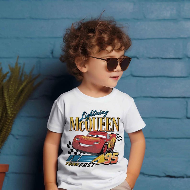 May include: White t-shirt featuring Lightning McQueen from the Cars movie. The graphic includes the car, the number 95, and the words "Lightning McQueen" and "Think Fast". The shirt is worn by a young child wearing sunglasses.