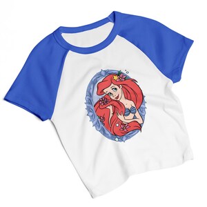May include: White and blue raglan t-shirt featuring a cartoon illustration of a red-haired mermaid in a blue oval frame. The mermaid is wearing a blue seashell bra and has pink flowers in her hair. The shirt has blue sleeves and a blue neckline.