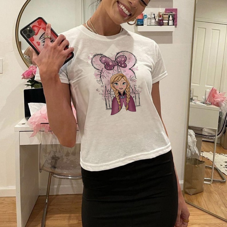 May include: A white short-sleeved t-shirt featuring a graphic print of a cartoon character with blonde braided hair, a purple cape, and a large purple polka-dot bow above the head, set against a background of a castle outline and small hearts.