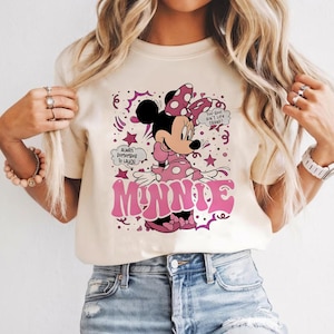 Op de afbeelding: Cr&egrave;mekleurig T-shirt met een afbeelding van Minnie Mouse in een roze jurk met stippen en een strik. Het woord "MINNIE" staat in roze, gestileerde letters. Tekstballonnen zeggen "Always Remember To Laugh" en "Too-Cool Ain't Life Grand?"